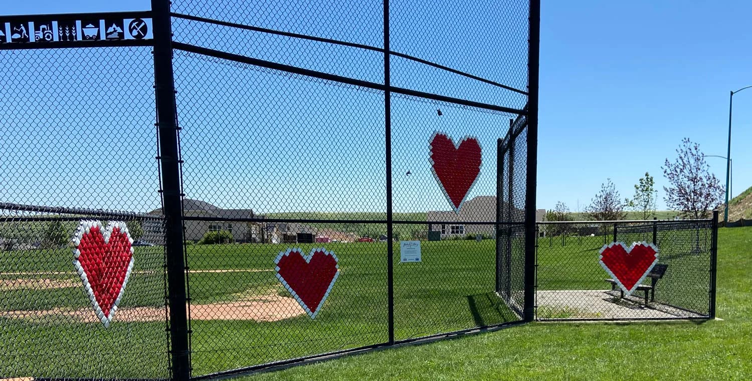 Red knitted or crocheted hearts installed on a chain-link fence surrounding a sports field.