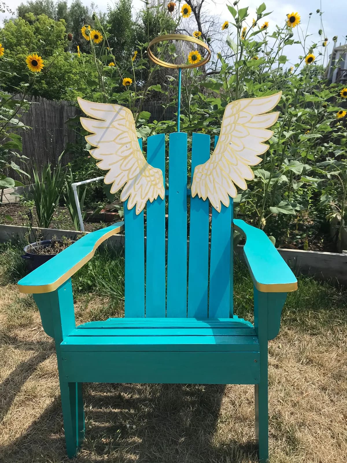 Turquoise wooden Adirondack chair with cream wings and gold halo set among blooming sunflowers in a garden installation.