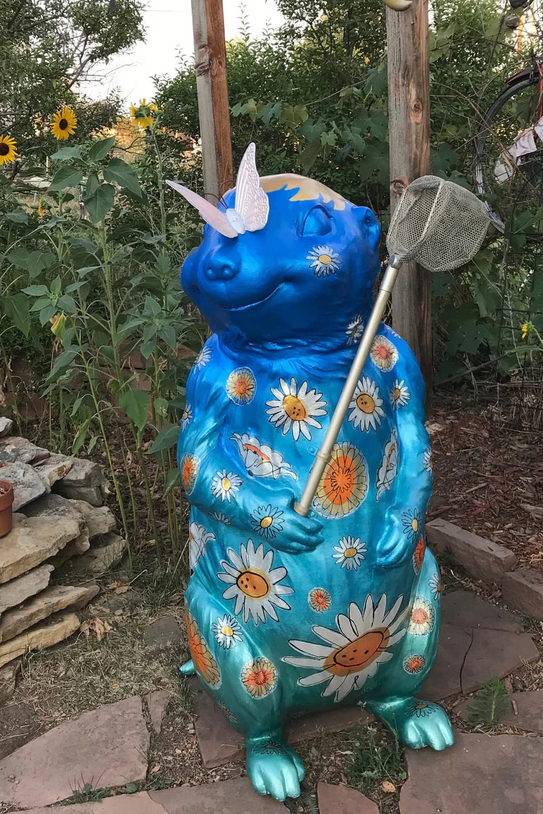 A blue metal sculpture of a smiling creature decorated with hand-painted sunflowers holds a butterfly net in a garden setting.