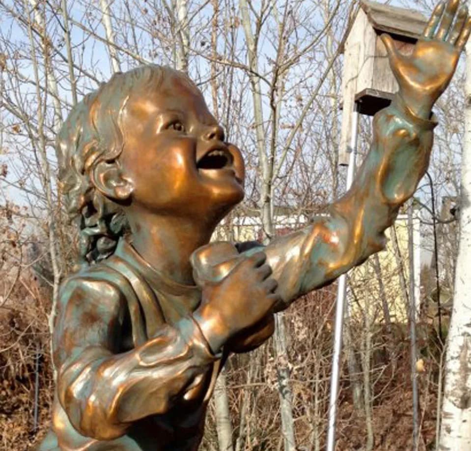 Bronze sculpture of a child figure reaching upward, displayed outdoors against bare trees.