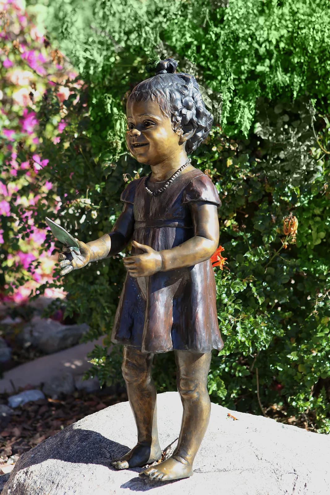 Bronze sculpture of a young girl in a dress holding flowers, set in a garden landscape.