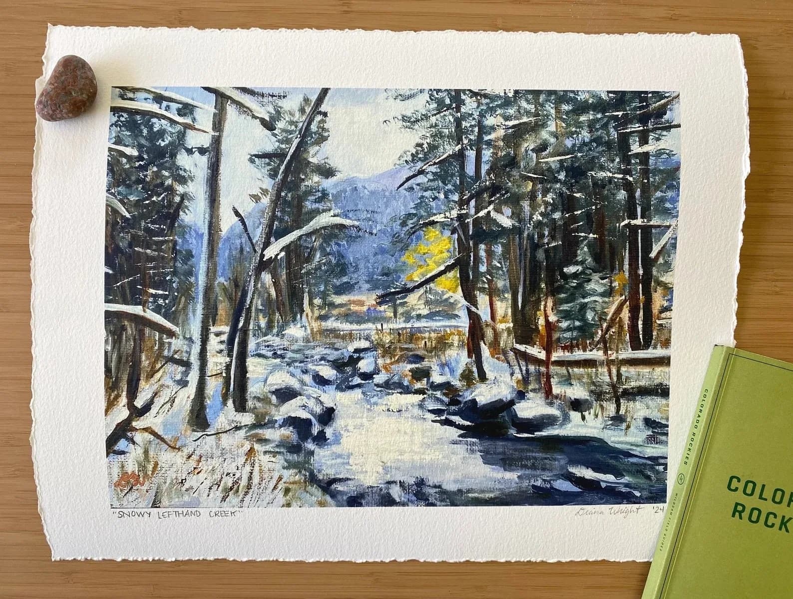 Watercolor landscape depicting a snowy winter forest opening toward the Boulder Flatirons beyond, with tall evergreens framing a frozen stream.