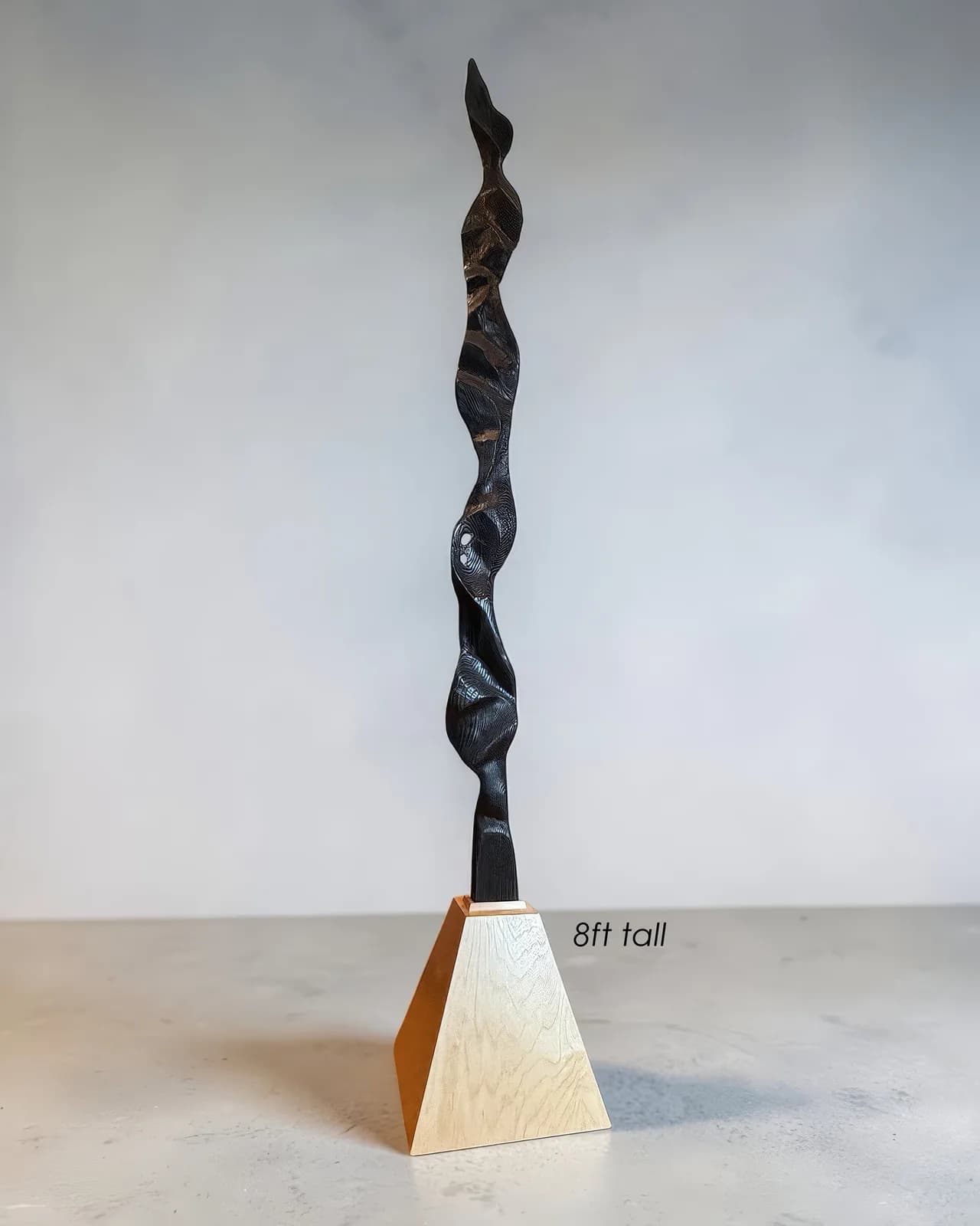 Tall twisted black wooden sculpture with a light wood base, approximately eight feet in height.
