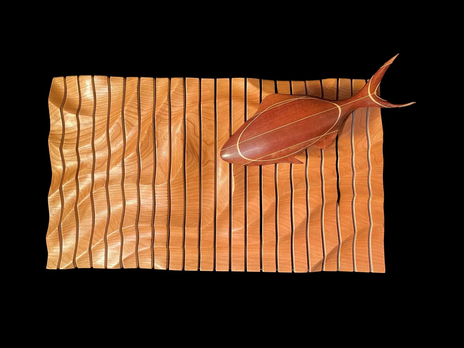 A carved wooden fish with detailed linear relief rests on a rippled wood panel against black background.