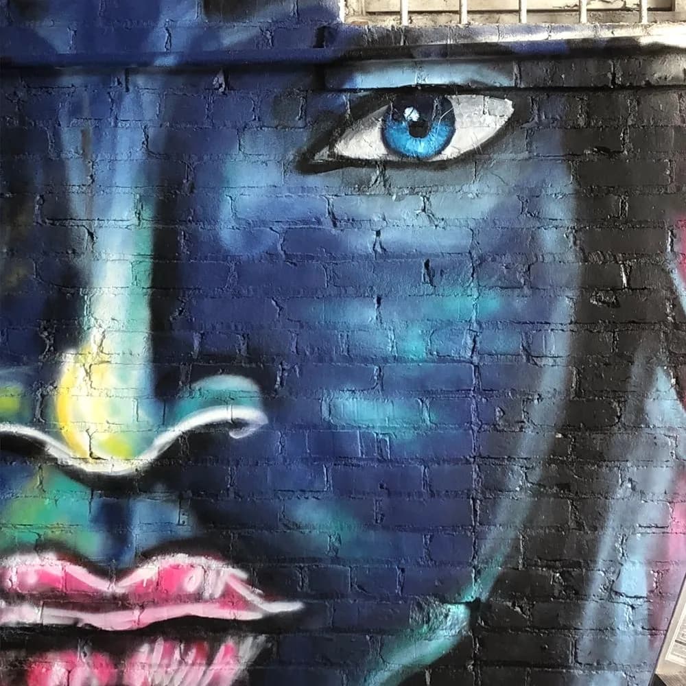 A stylized blue face with striking cyan eye and pink lips rendered in spray paint on brick.