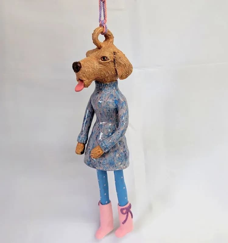 A whimsical ceramic dog figure in blue dress and pink boots, with cork head and hanging string.