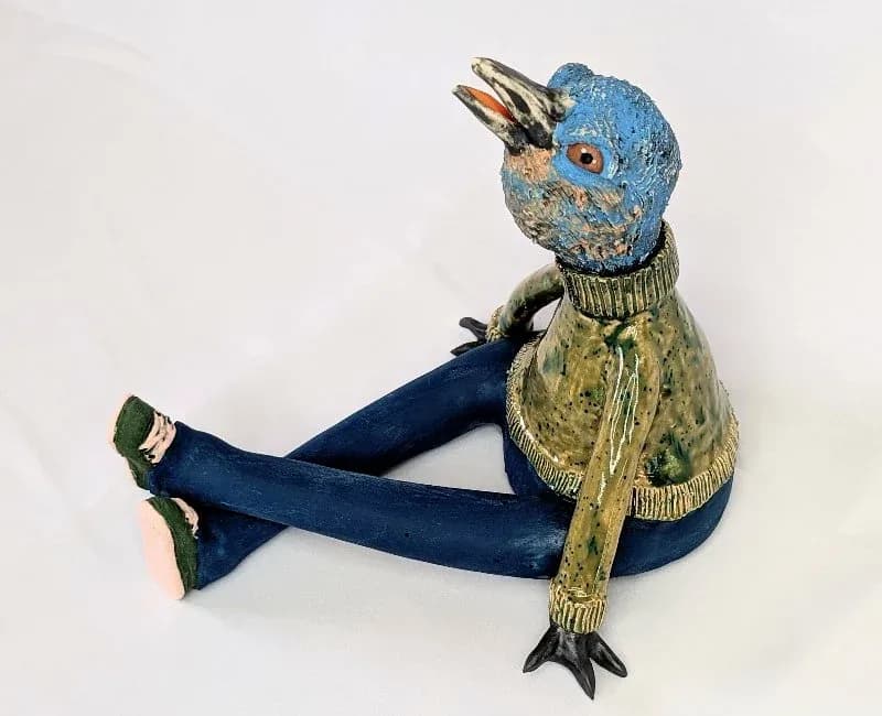 Whimsical ceramic bird figure with blue head, tan coat, and dark blue trousers, photographed against white.