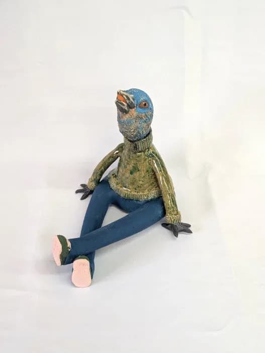 A whimsical ceramic bird figure with outstretched blue fabric legs wears a golden collar detail.
