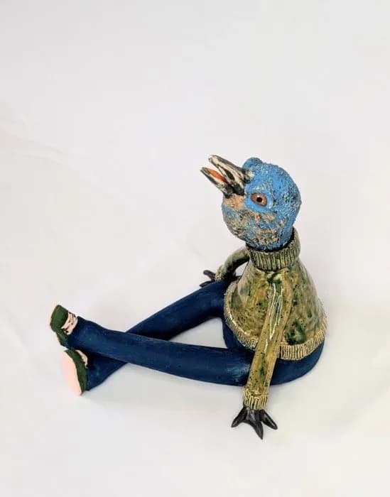 Handmade ceramic bird sculpture with blue head, yellow jacket, and navy legs posed against white background.