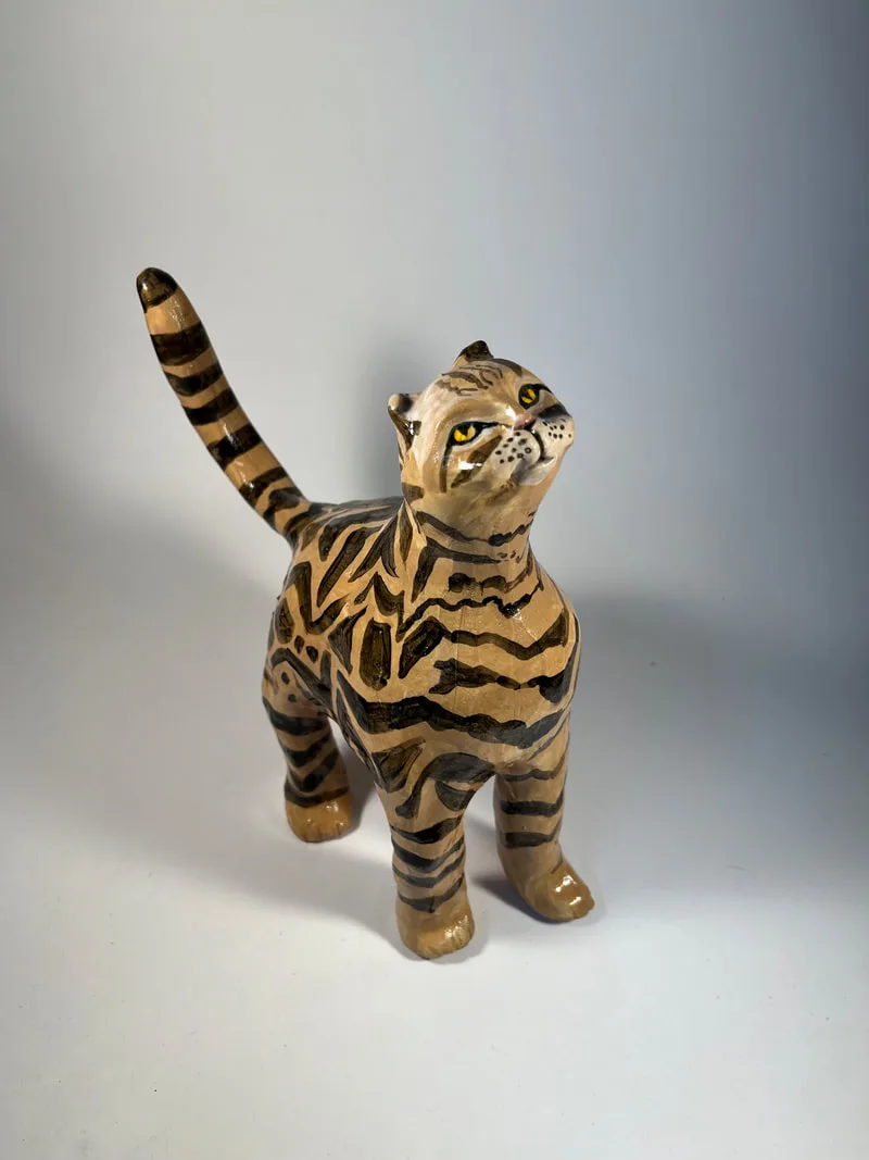 A glazed ceramic cat figure with black stripes, painted eyes, and raised tail, rendered in warm earthtone tones.