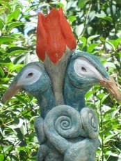 Abstract ceramic sculpture with spiraled blue-gray body and red flower crown, set in garden foliage.