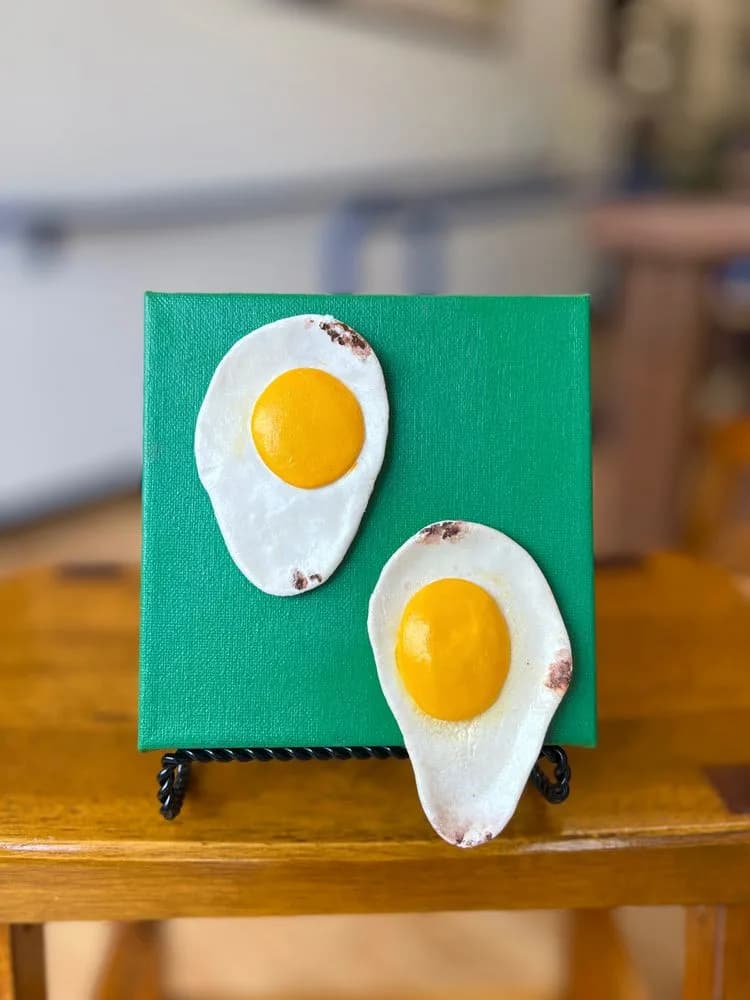 Two fried eggs with dimensional white shells protrude from a bright green painted canvas on a wooden shelf.