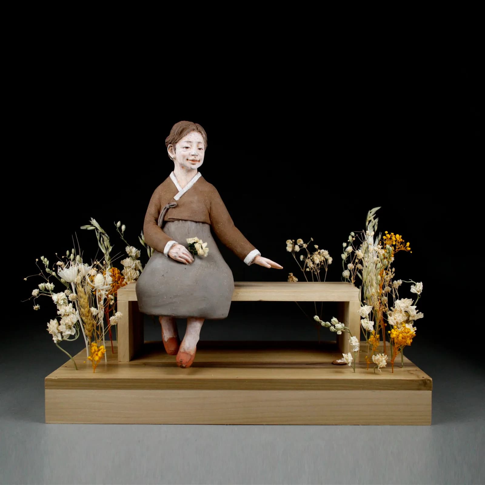 A carved wooden figure sits on a bench surrounded by dried flowers and grasses, rendered in warm earth tones.