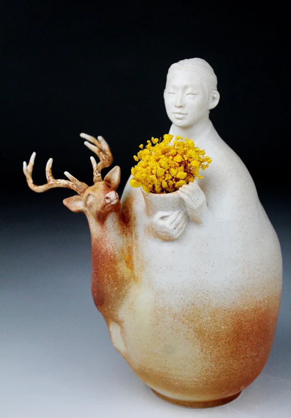 A ceramic vessel with a human head and torso form holds yellow flowers beside a cast deer head with antlers.