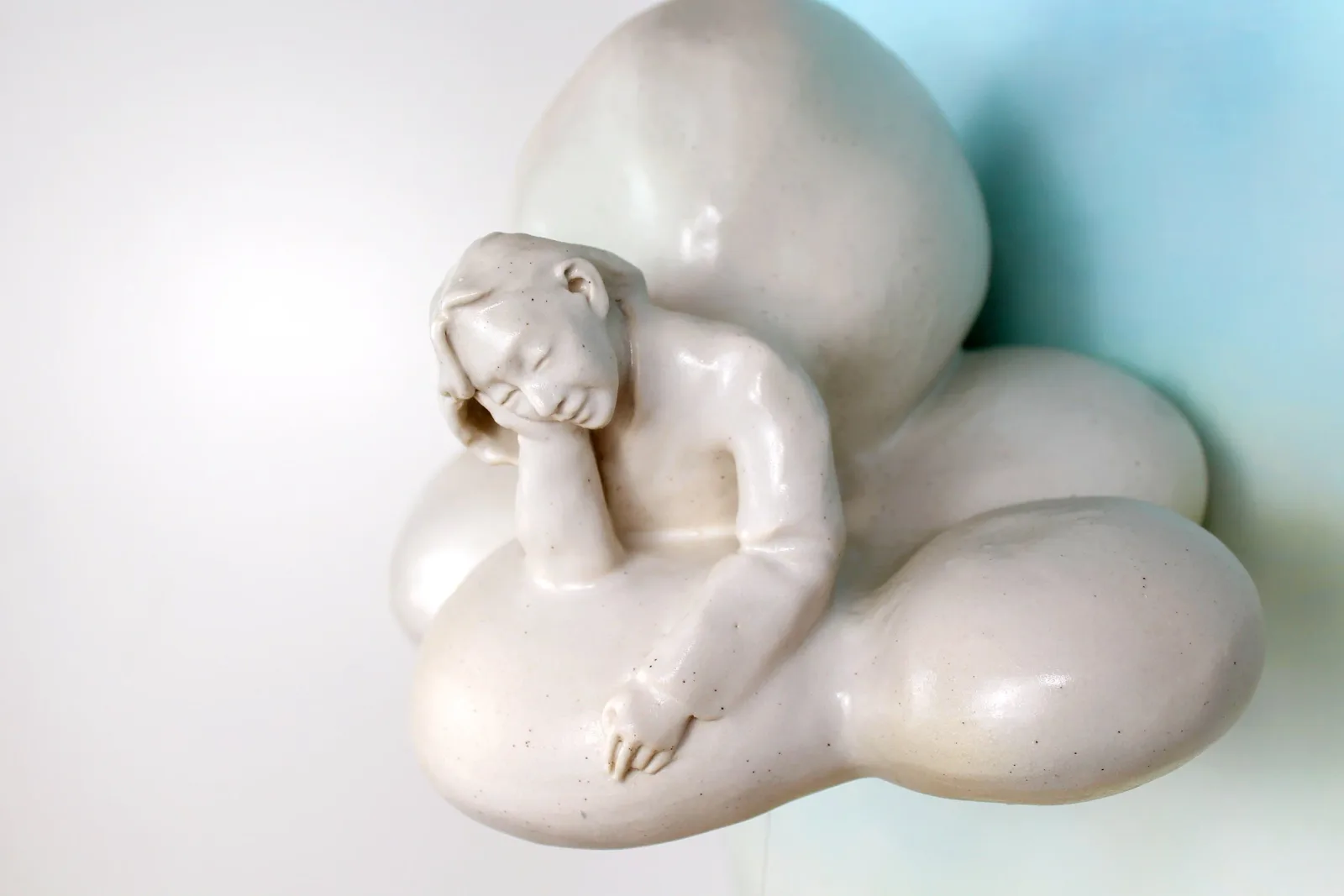 A white ceramic figurative sculpture of a reclining child nestled among organic ovoid forms.