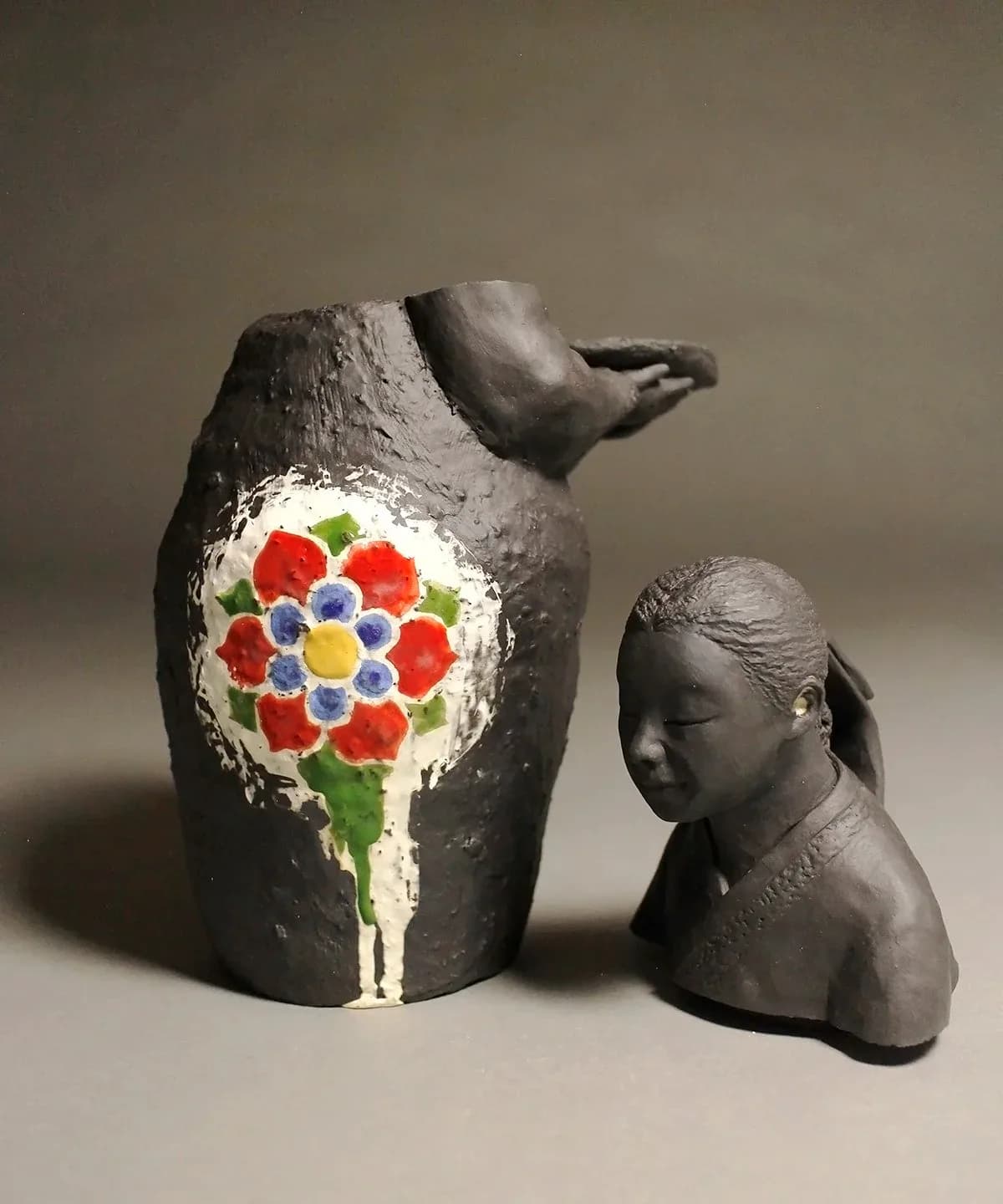 Two hand-built ceramic vessels in clay with a sculptural head and a pitcher form decorated with a bold red-and-blue floral motif.