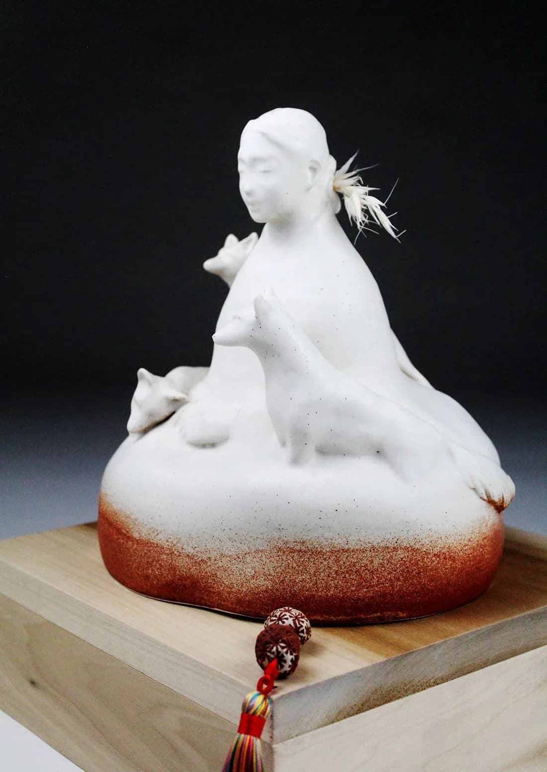 Seated ceramic figurative sculpture with white glaze, feather detail, and decorative tassel ornament.