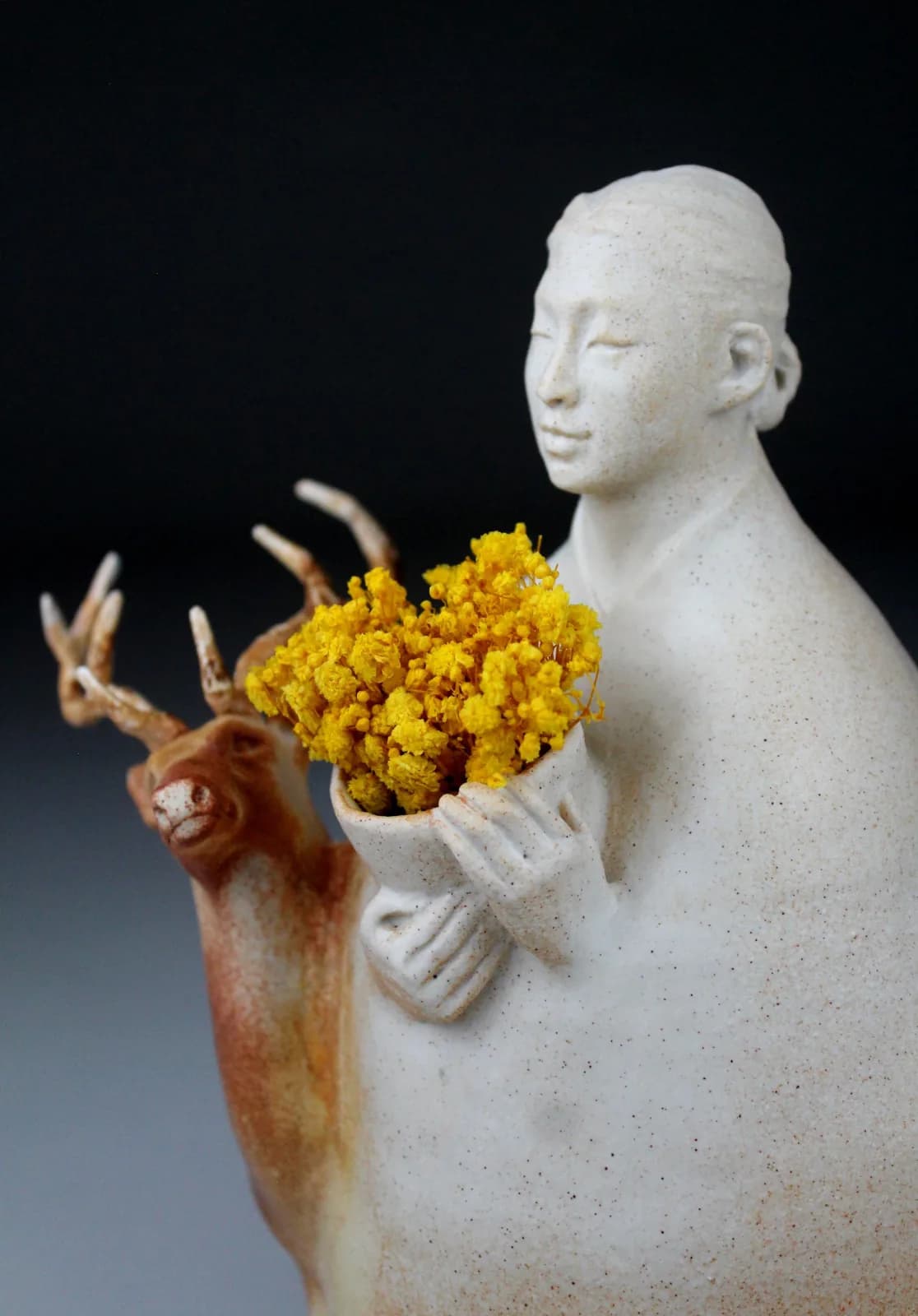 Ceramic figure holding a deer with yellow flowers against a dark background.