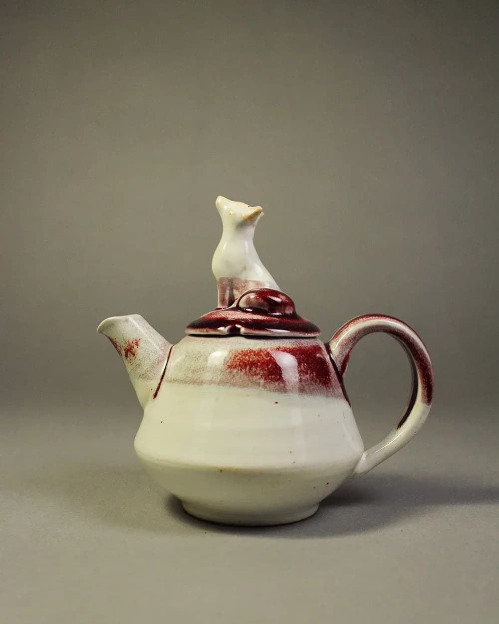 Ceramic teapot with cream-and-burgundy glaze, featuring a sculptural bird finial on the lid.