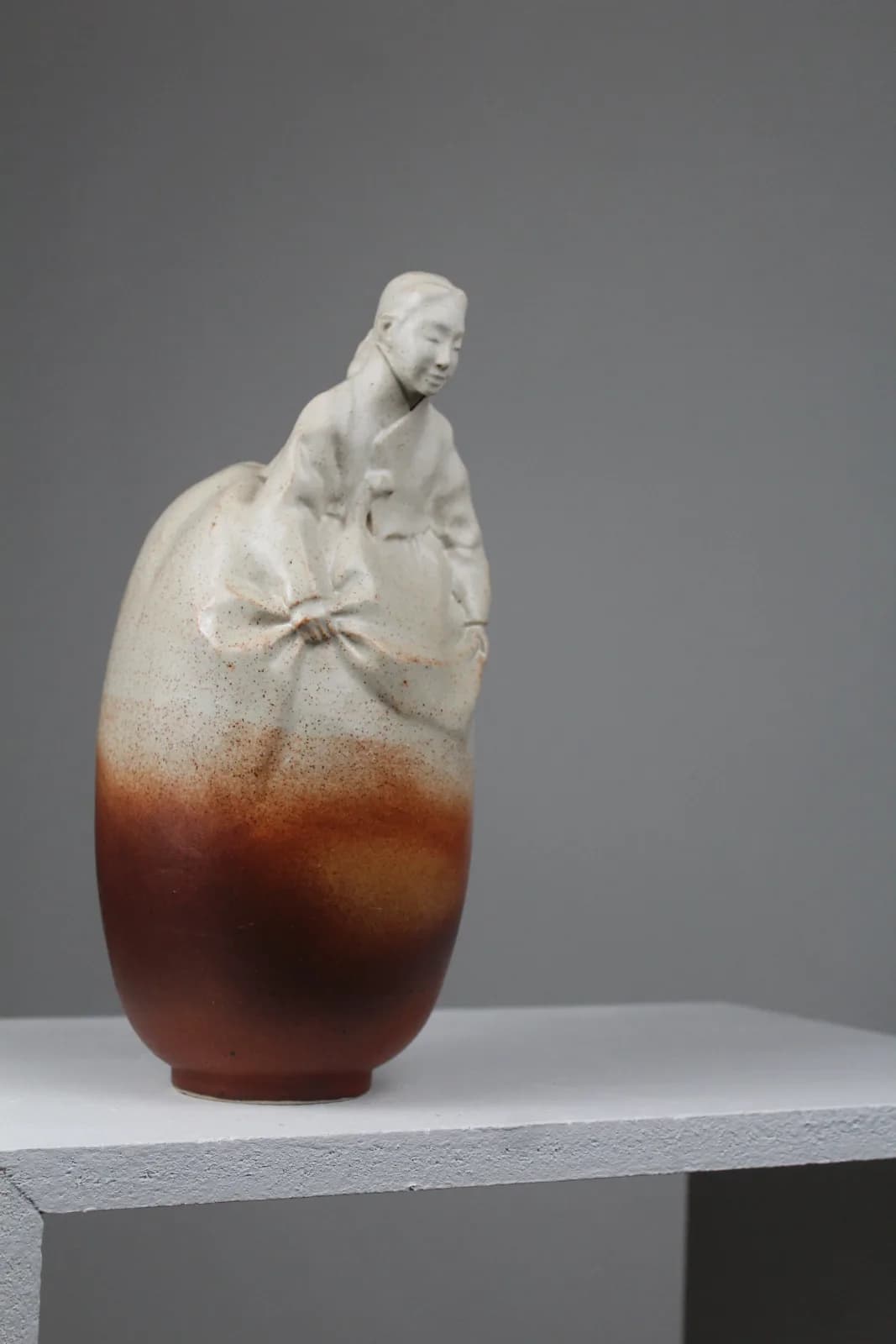 A ceramic vessel depicting a seated figure emerging from a rust-glazed egg form in stoneware.