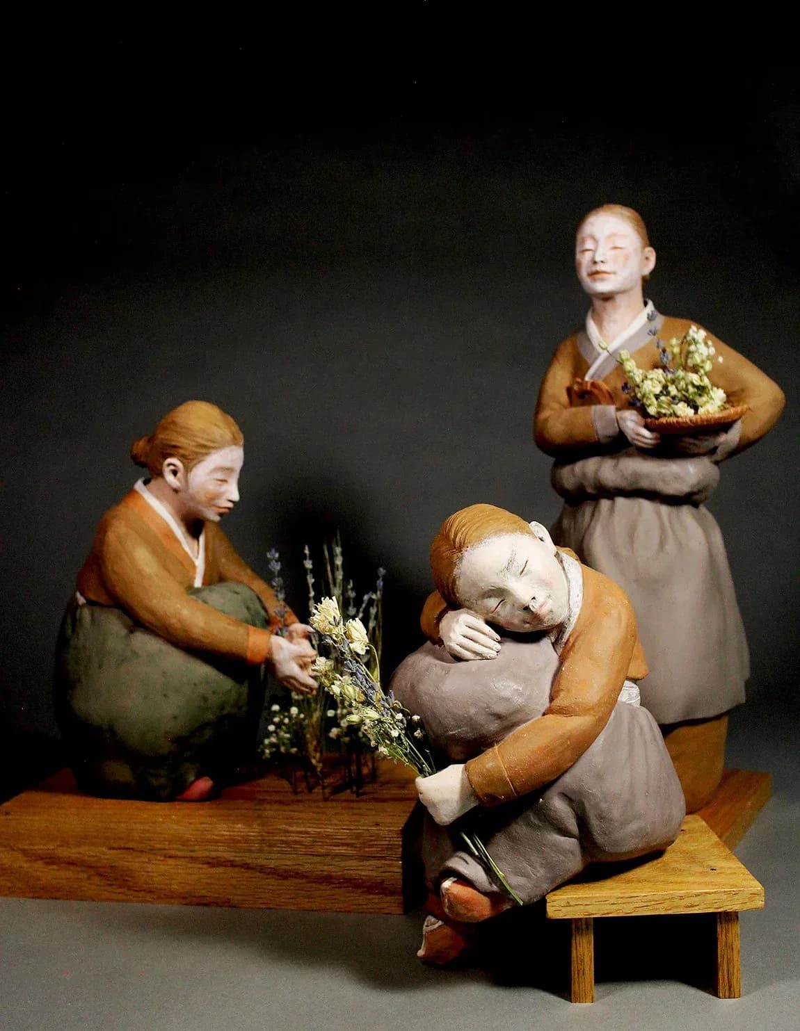 Three ceramic figures with earth-toned clothing seated with dried flowers on a wooden base.