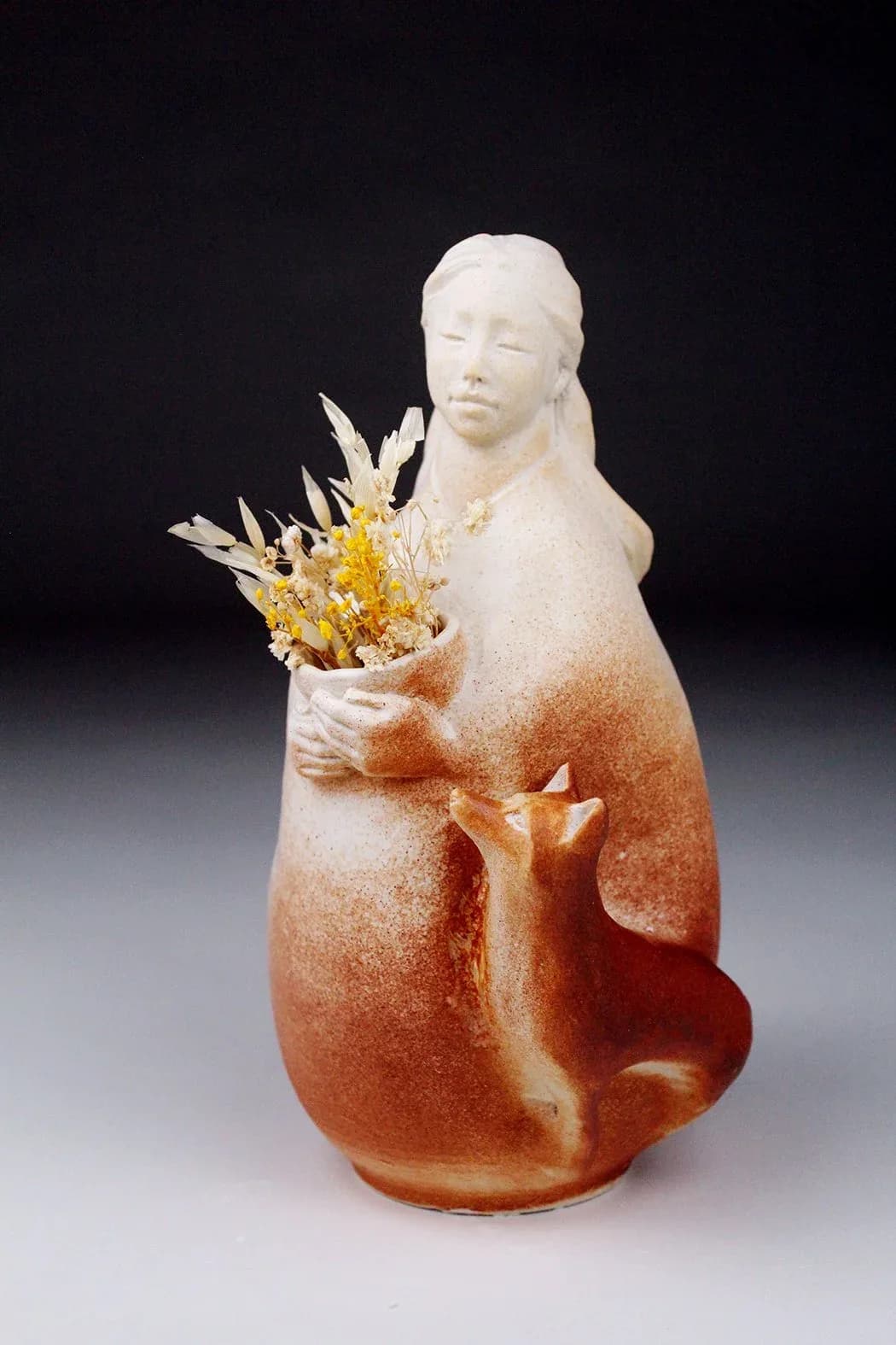 Ceramic figurative sculpture of a woman holding dried flowers, with gradient glazing from white to warm gold and rust tones.