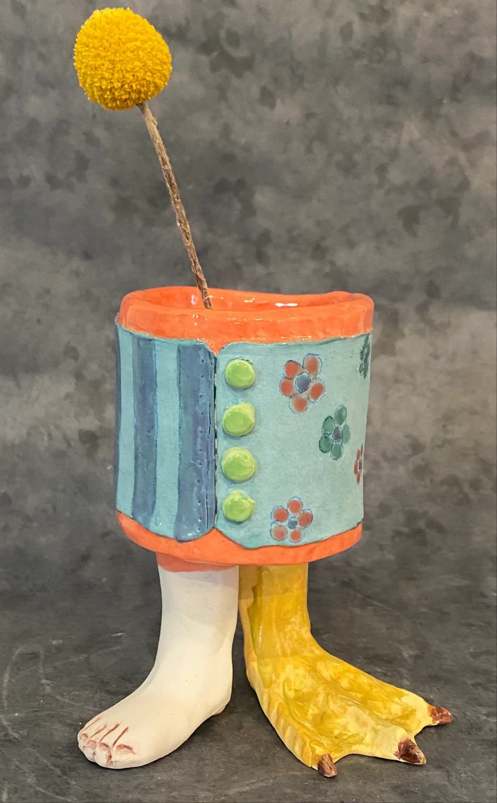 Whimsical ceramic sculpture of a creature with a yellow pompom head, patterned torso, and bird-like feet.