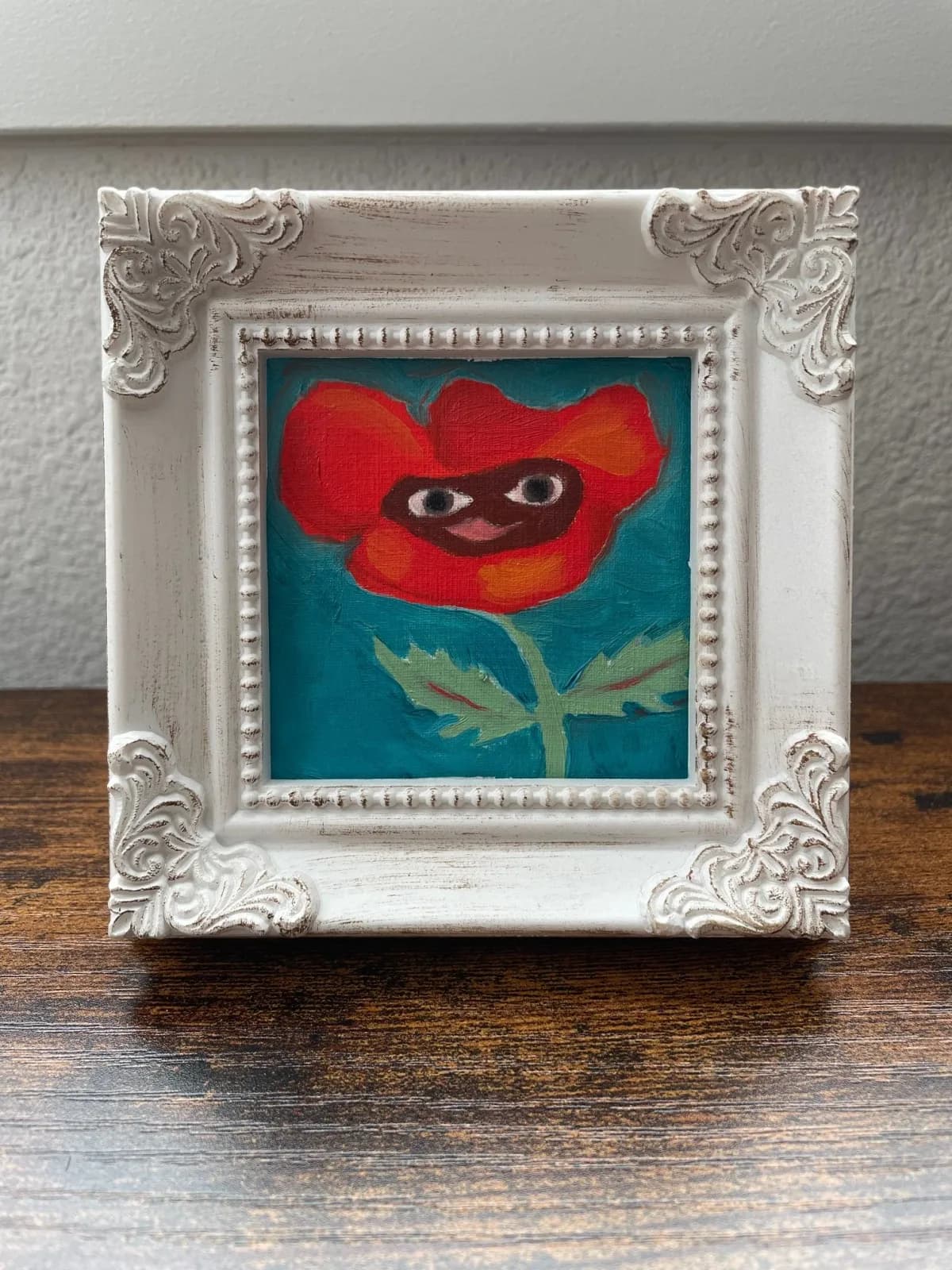 A red poppy with expressive eyes painted in acrylic within an ornate white frame.