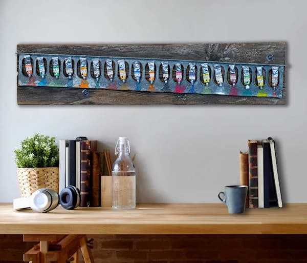 A horizontal mixed-media assembly on reclaimed wood featuring a row of miniature painted objects in vivid hues.
