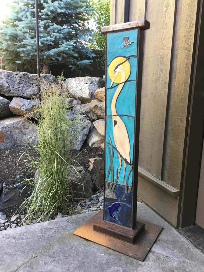 A stained-glass heron stands on a wooden base, depicting the bird in blue water with yellow sun overhead.