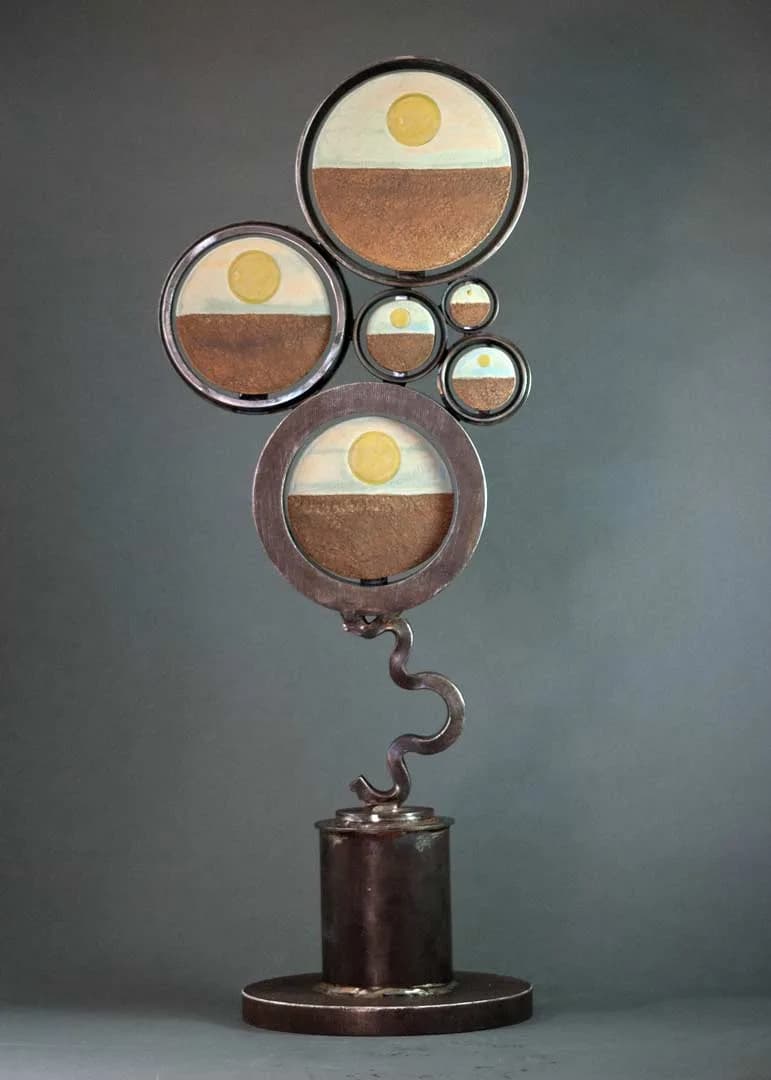 A bronze kinetic sculpture with stacked spheres containing sand and yellow orbs, balanced on a curved stem.