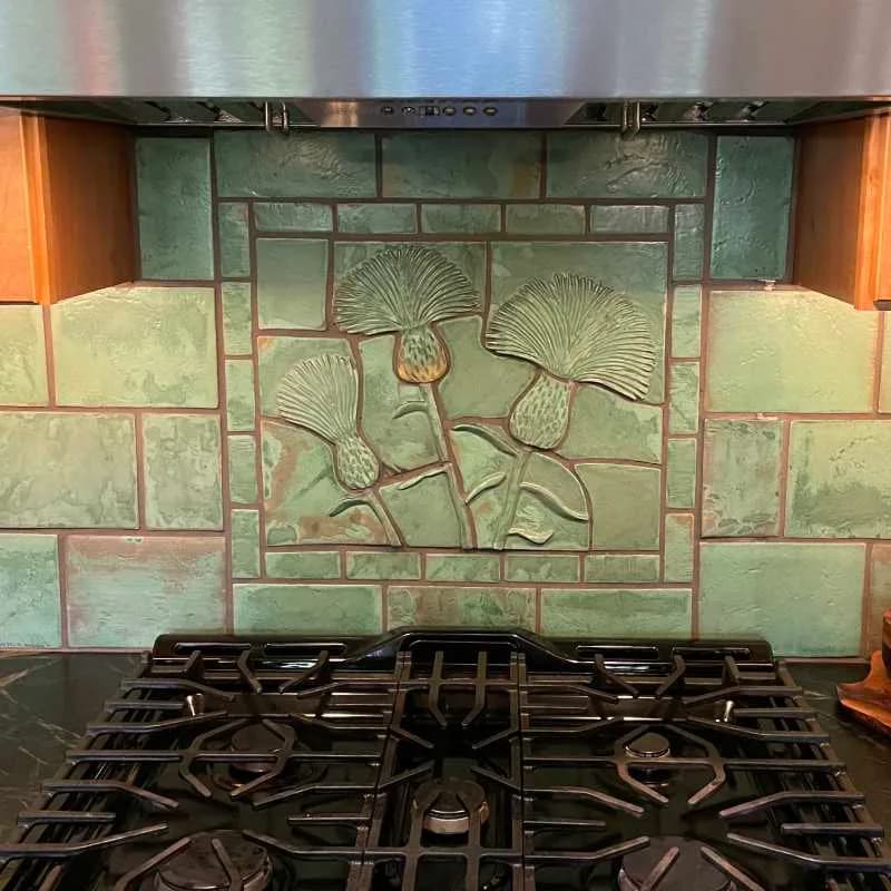 Ceramic tile backsplash with embossed ginkgo leaf design in green and cream tones, installed above a gas cooktop.