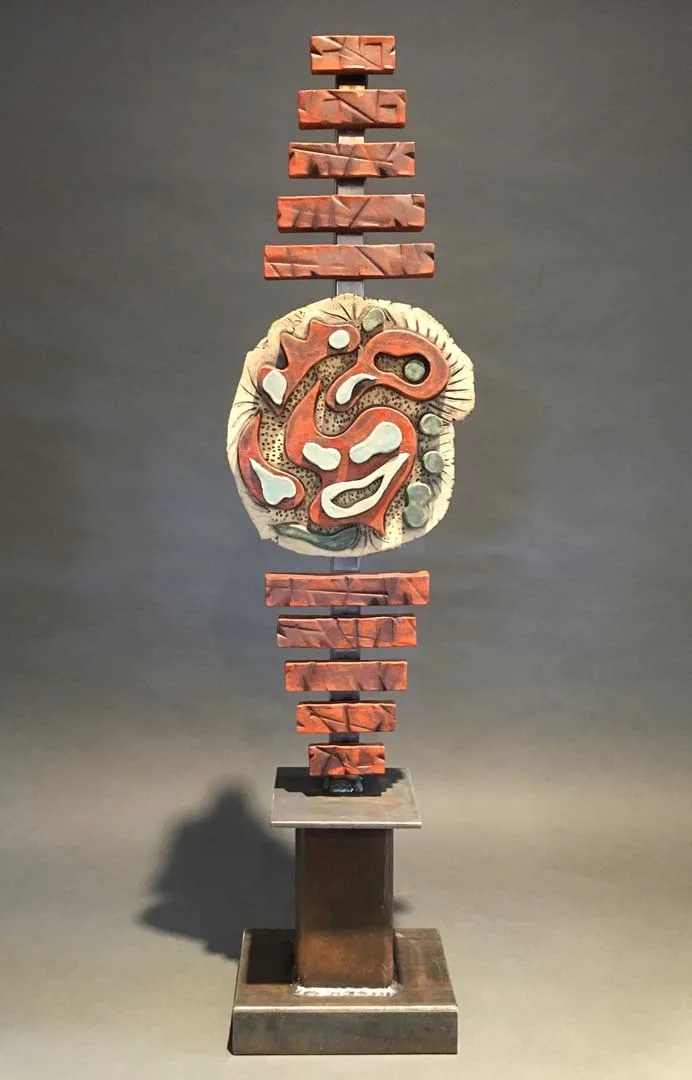 Mixed media sculpture combining a ceramic mask with stacked and balanced brick segments on a metal base.