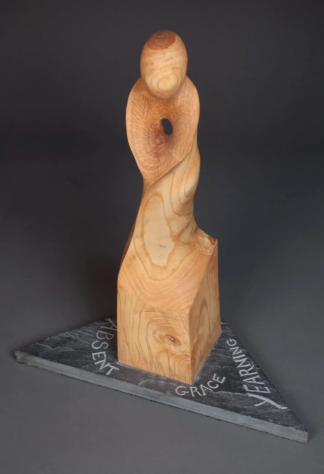 Abstract wood-carved figure with an engraved slate base inscribed with words.