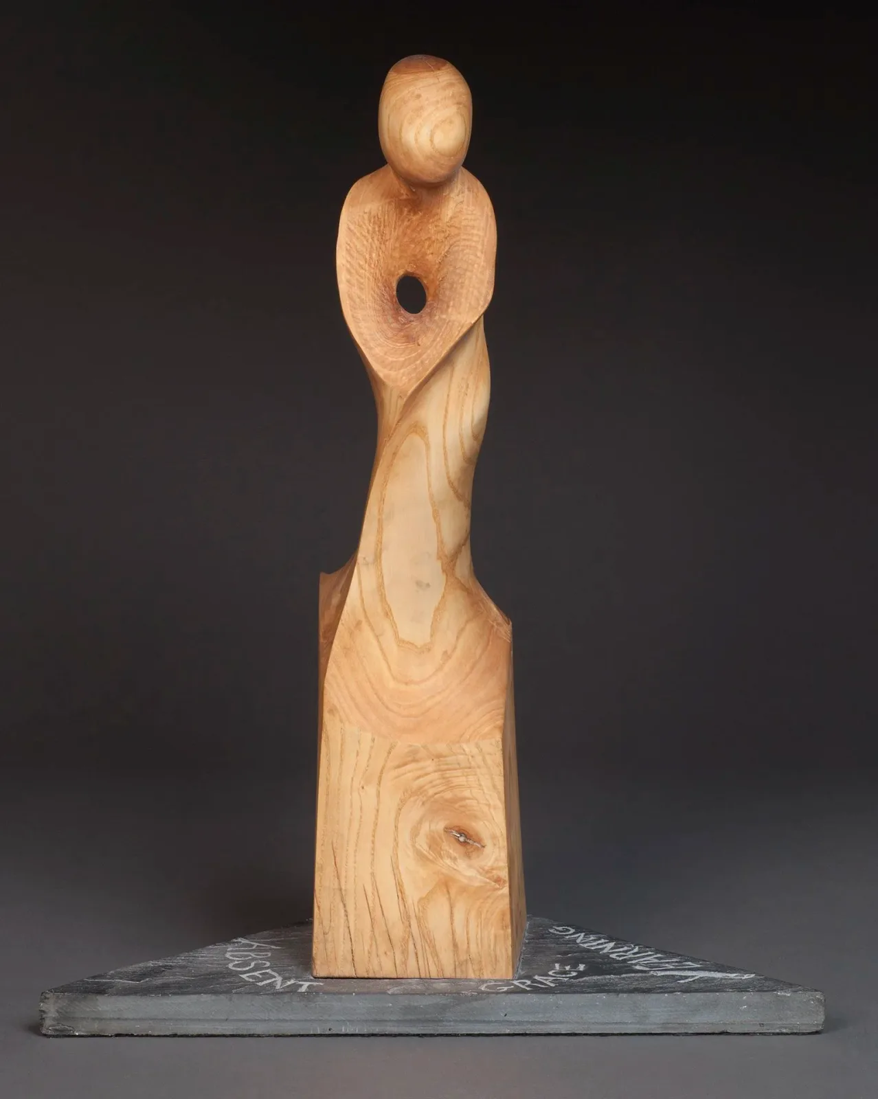 Abstract wooden figure carved from light wood, mounted on a dark rectangular base.