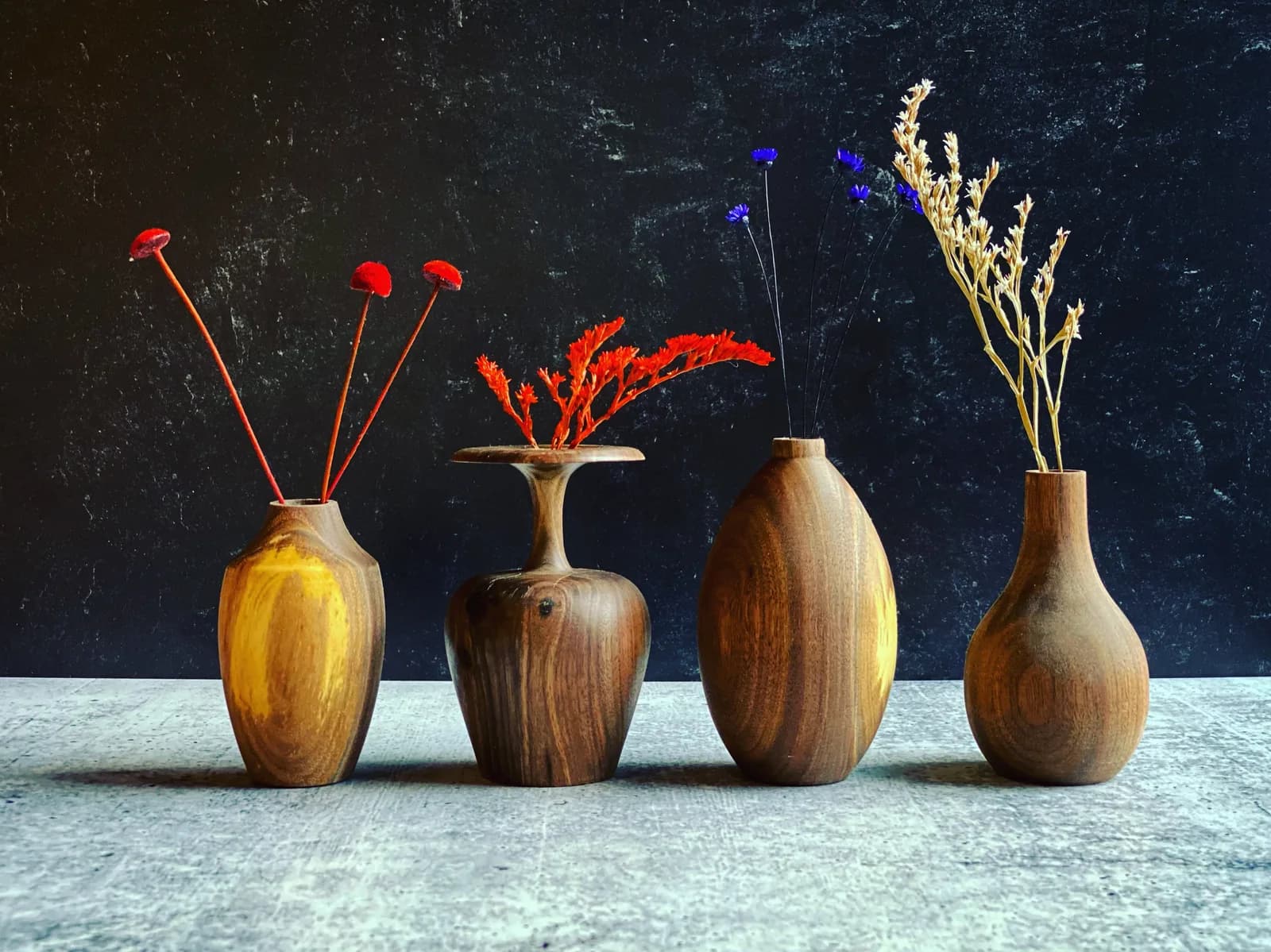 Four wooden vases hold dried and fresh flowers against a dark textured background in oil paint.