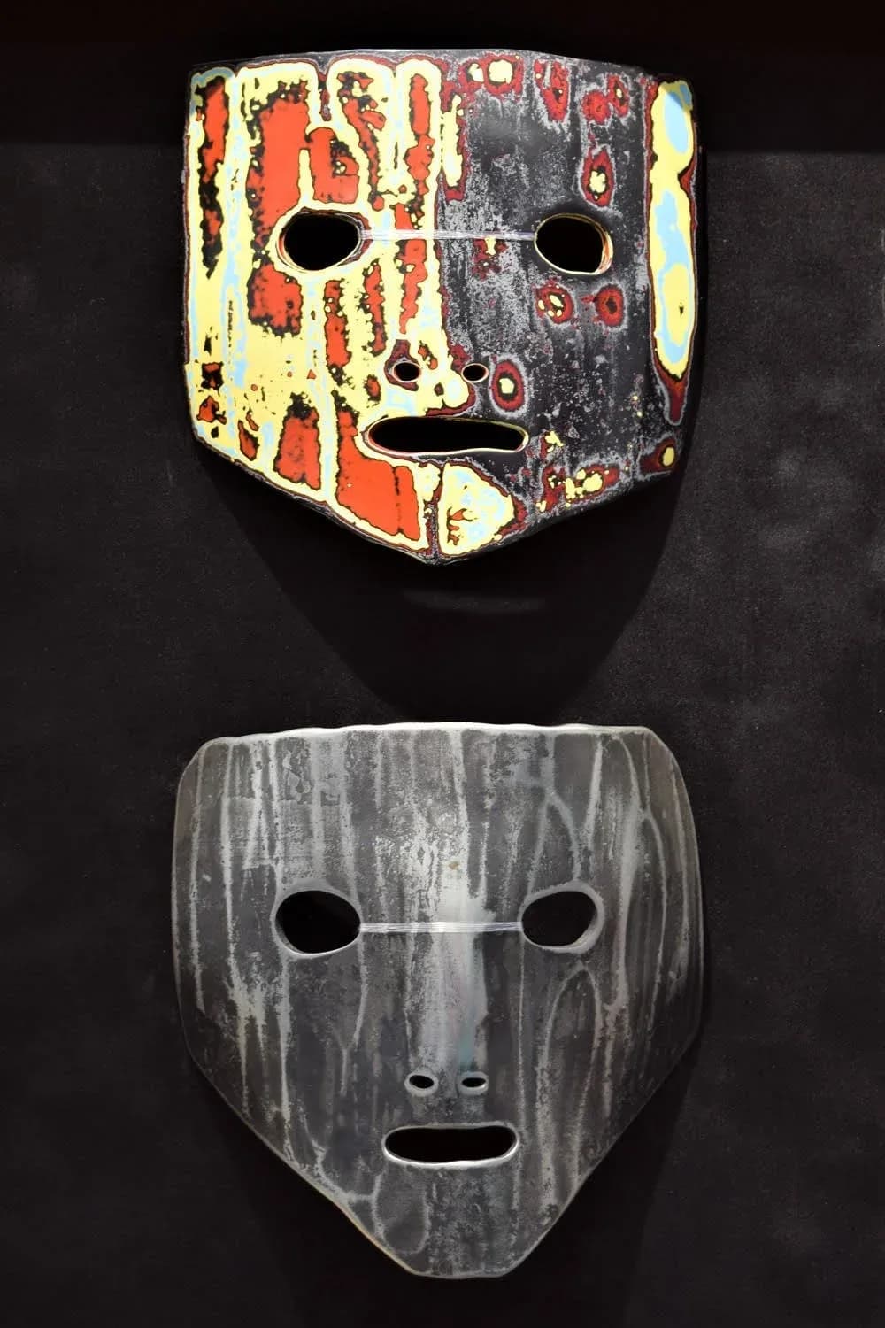 Two clay masks with stylized faces; upper mask in flame pea, cream, and charcoal; lower mask in tundora gray with subtle texture.