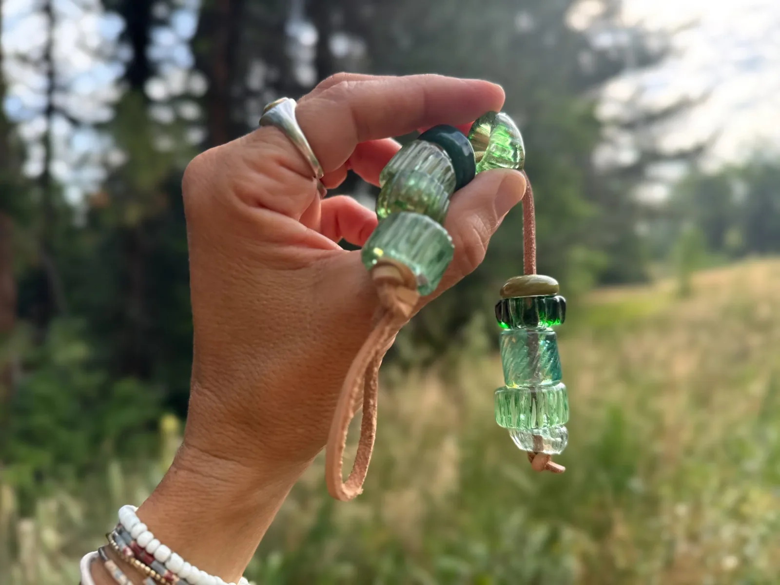 Handmade glass bead pendant with green and kelp-toned beads suspended from a cord against Boulder Flatirons backdrop.