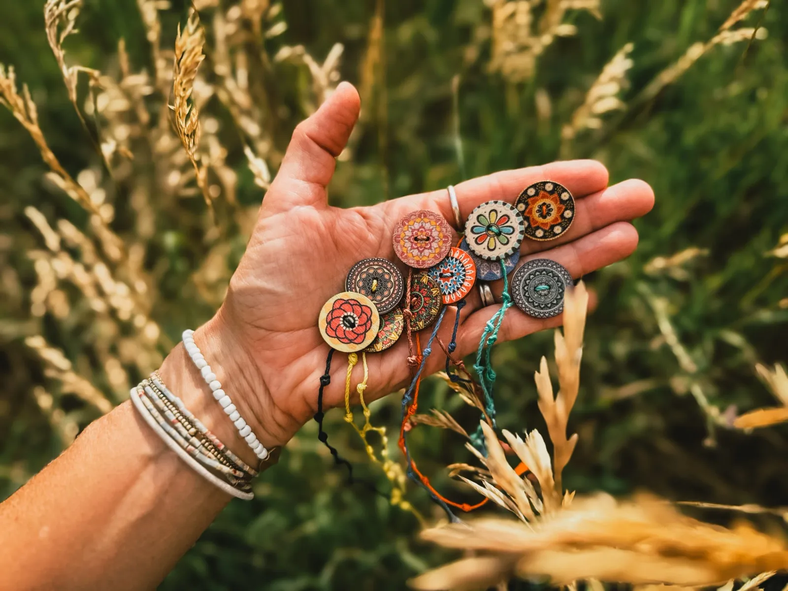 Hand-painted metal and ceramic medallion pins with twine strands displayed in a palm against dried grass.