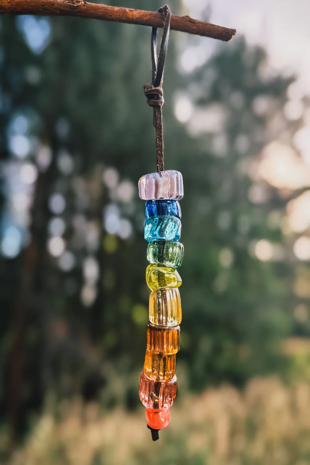 Stacked glass pendant in gradient rainbow colors suspended from a cord on wood branch.