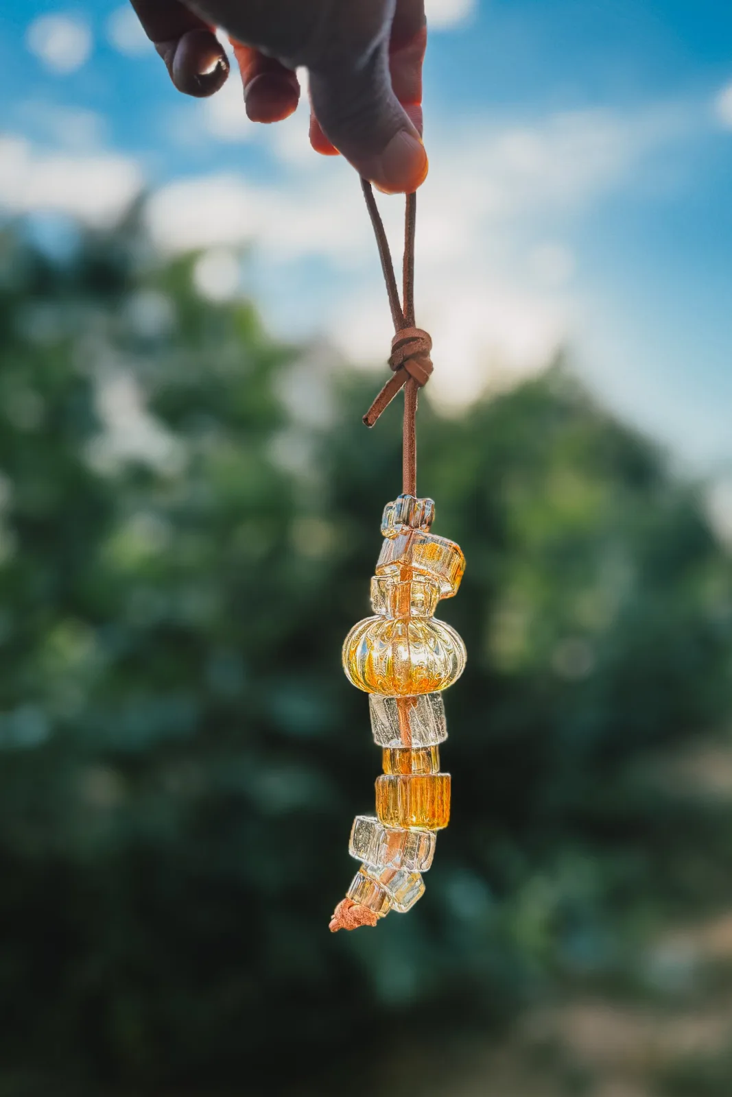 Amber and clear glass beads strung on leather cord, suspended against a blurred green and blue outdoor backdrop.