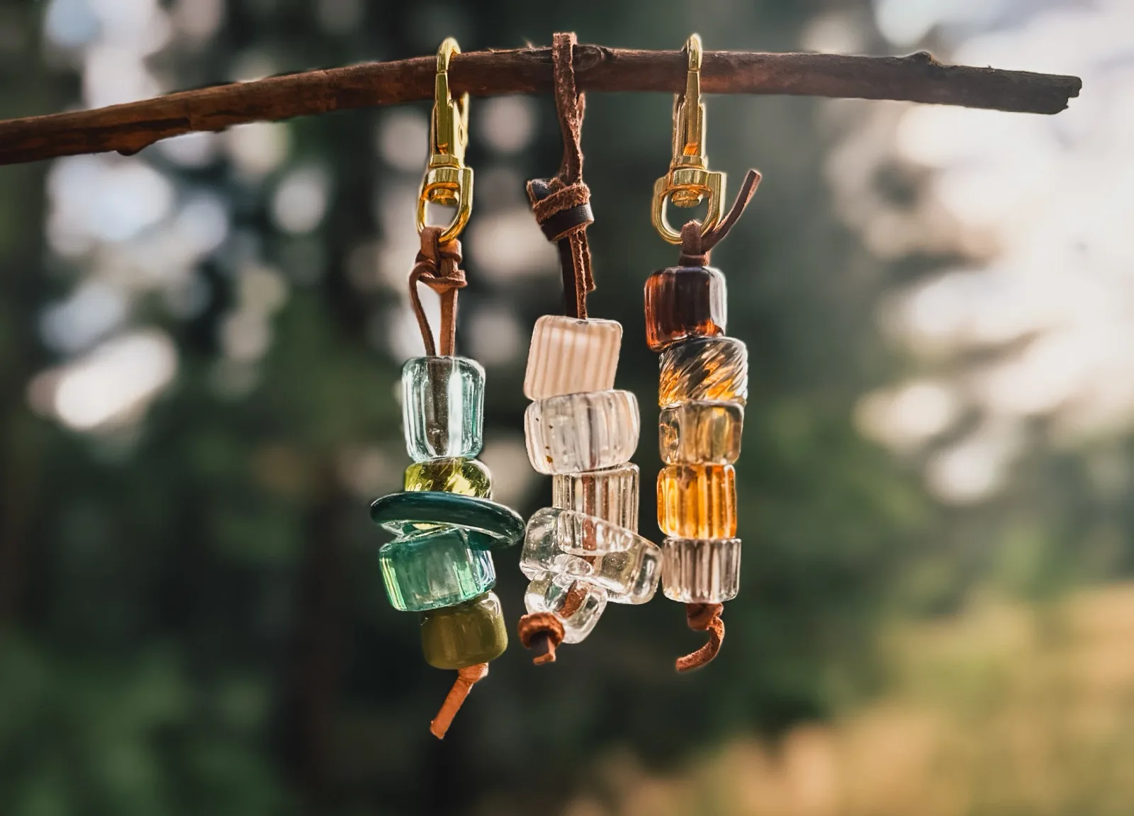Three beaded keychains with glass cylinders and brass hardware suspended from a weathered wood branch.