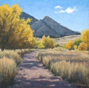 A dirt trail winds through golden aspens toward the Boulder Flatirons under pale blue sky.