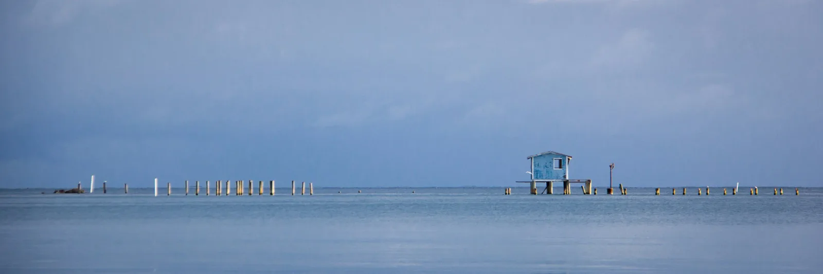 A minimalist coastal landscape with a distant pier and lift station rendered in shades of polo blue and kashmir blue.
