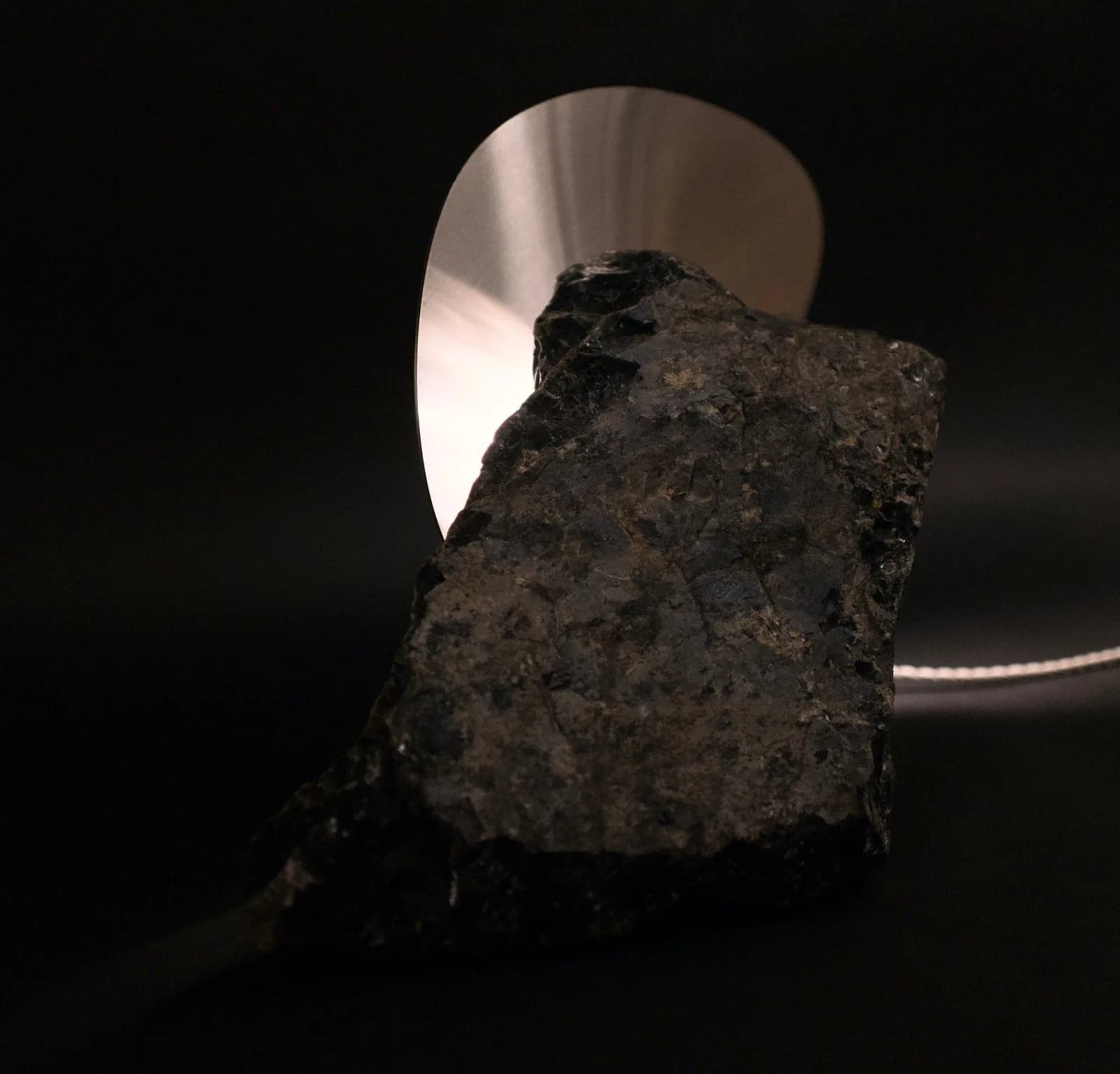 Raw stone form backlit by a luminous metallic disc against deep shadow.
